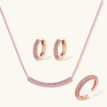 Rose gold jewelry set with pavé cubic zirconia stones including curved necklace, hoop earrings, and adjustable ring on cream background.