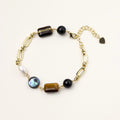 Gold chain bracelet with tiger's eye, obsidian, pearl, and iridescent abalone stones on cream background.