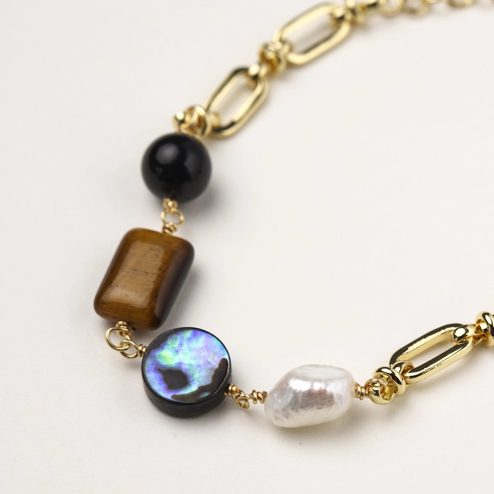 Gold chain bracelet with tiger's eye, obsidian, pearl, and iridescent abalone stones on cream background.