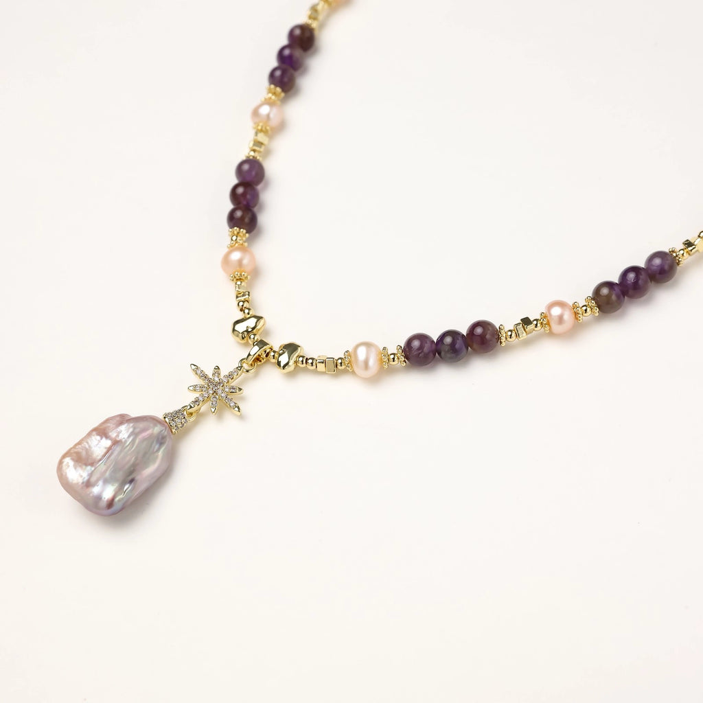 Gold necklace with iridescent baroque pearl pendant, purple amethyst beads, and crystal star charm on cream background