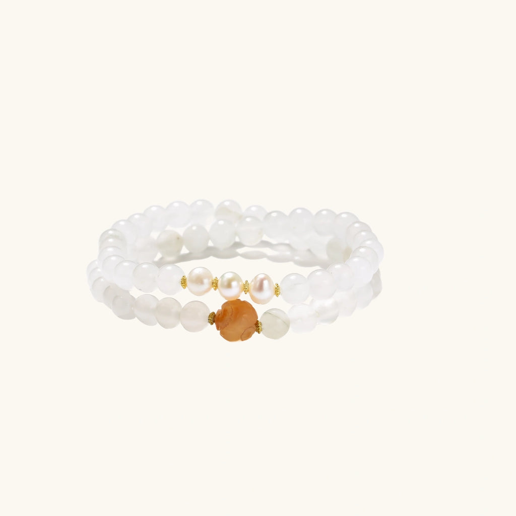 Three layered white beaded bracelets with carved orange agate lotus centerpiece and freshwater pearls on cream background.