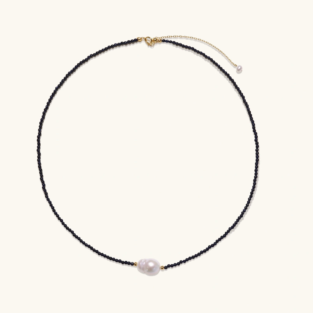 Delicate necklace with black blue sandstone beads and baroque pearl center on white background with gold accents.