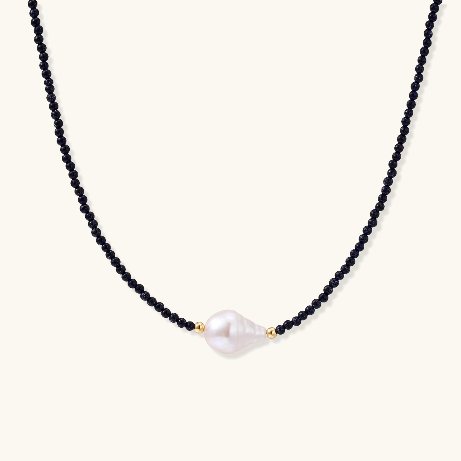 Delicate necklace with black blue sandstone beads and baroque pearl center on white background with gold accents.