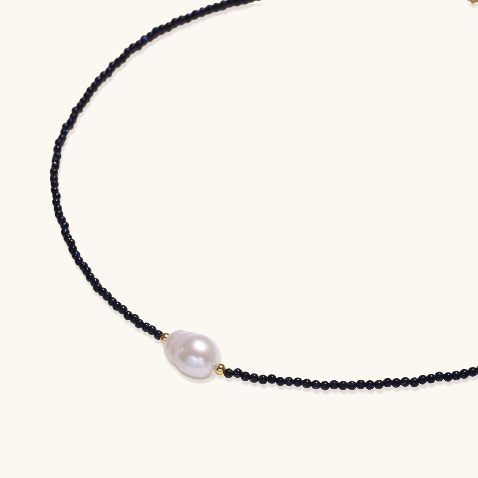 Delicate necklace with black blue sandstone beads and baroque pearl center on white background with gold accents.