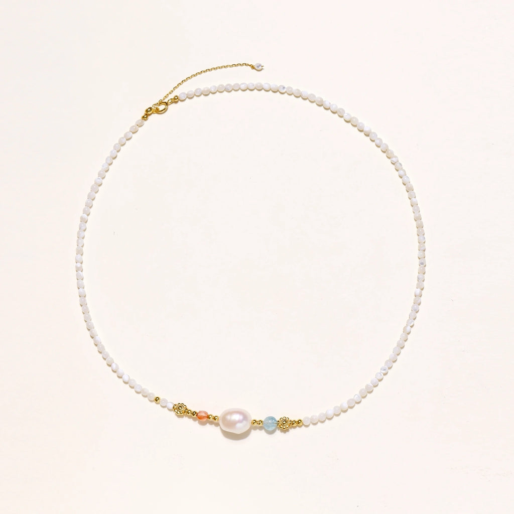 Delicate white beaded necklace with baroque pearl center, gold accents, and coral and aqua gemstones on cream background.