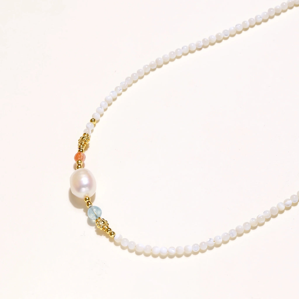 Delicate white beaded necklace with baroque pearl center, gold accents, and coral and aqua gemstones on cream background.