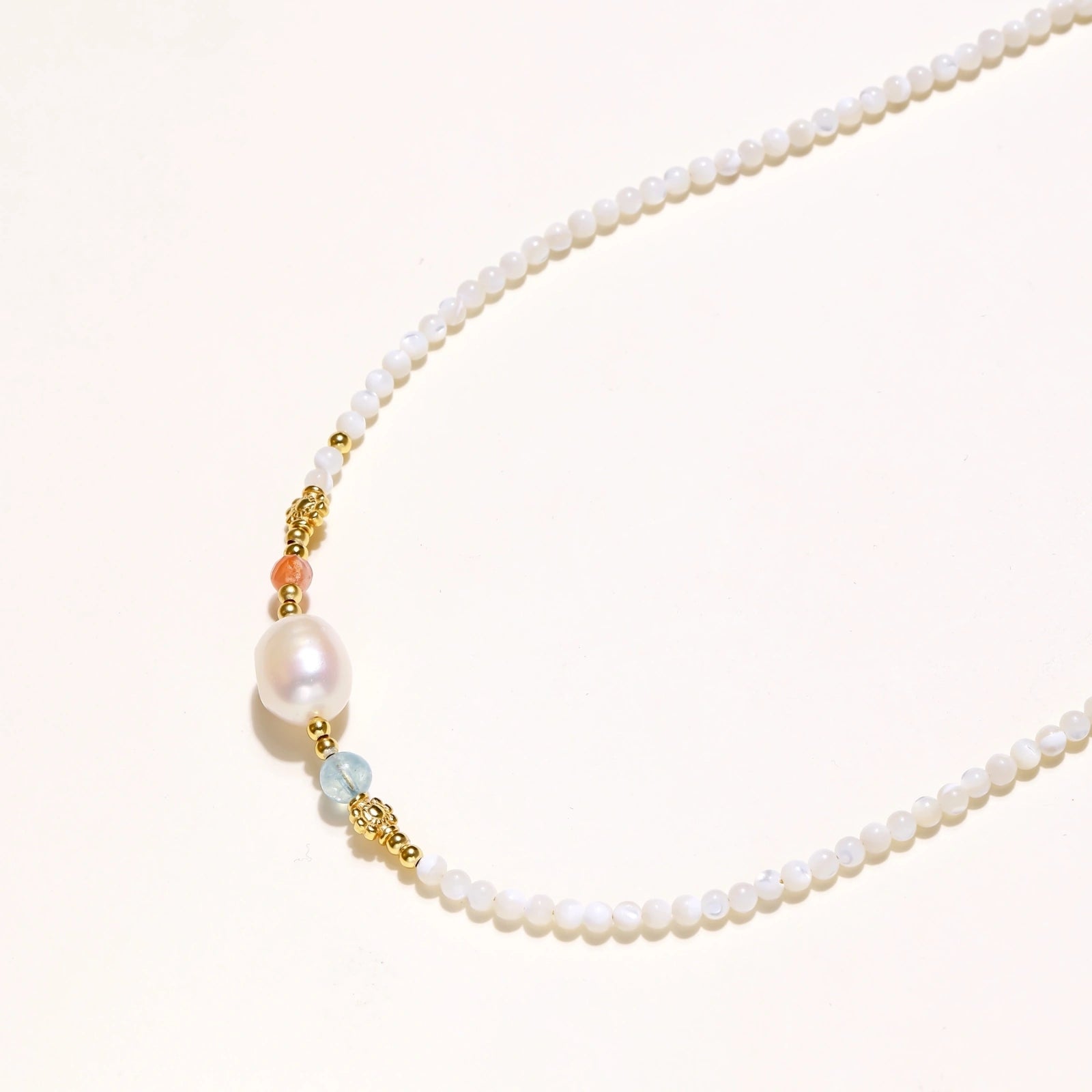 Delicate white beaded necklace with baroque pearl center, gold accents, and coral and aqua gemstones on cream background.