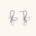 Silver bow-shaped stud earrings with baguette crystals and dangling white freshwater pearls on cream background