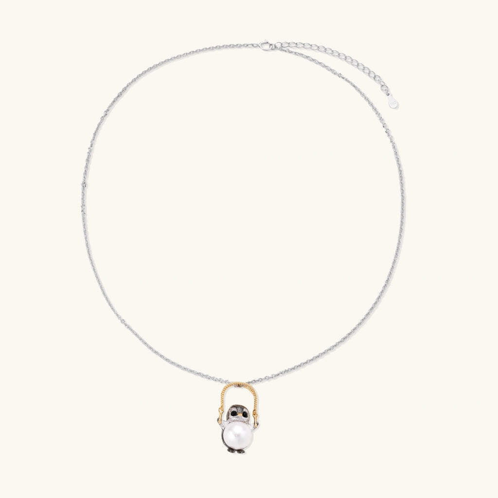 Whimsical owl pendant holding a white pearl on a gold swing, rhodium-plated silver chain on cream background.