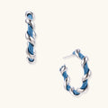 White gold-plated hoops intertwined with blue denim fabric in a twisted rope design on cream background.