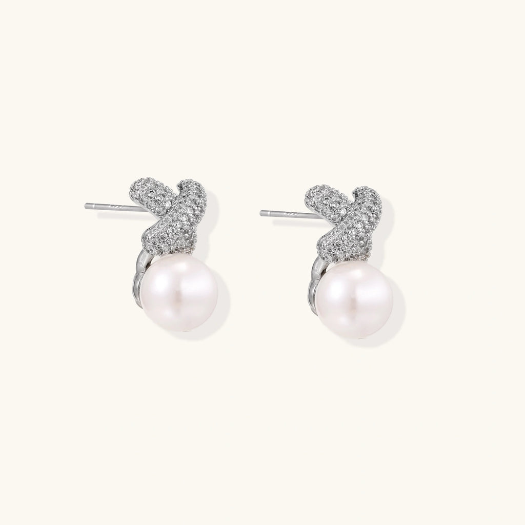 White gold plated studs featuring pavé X-shaped tops with freshwater pearl drops on cream background.