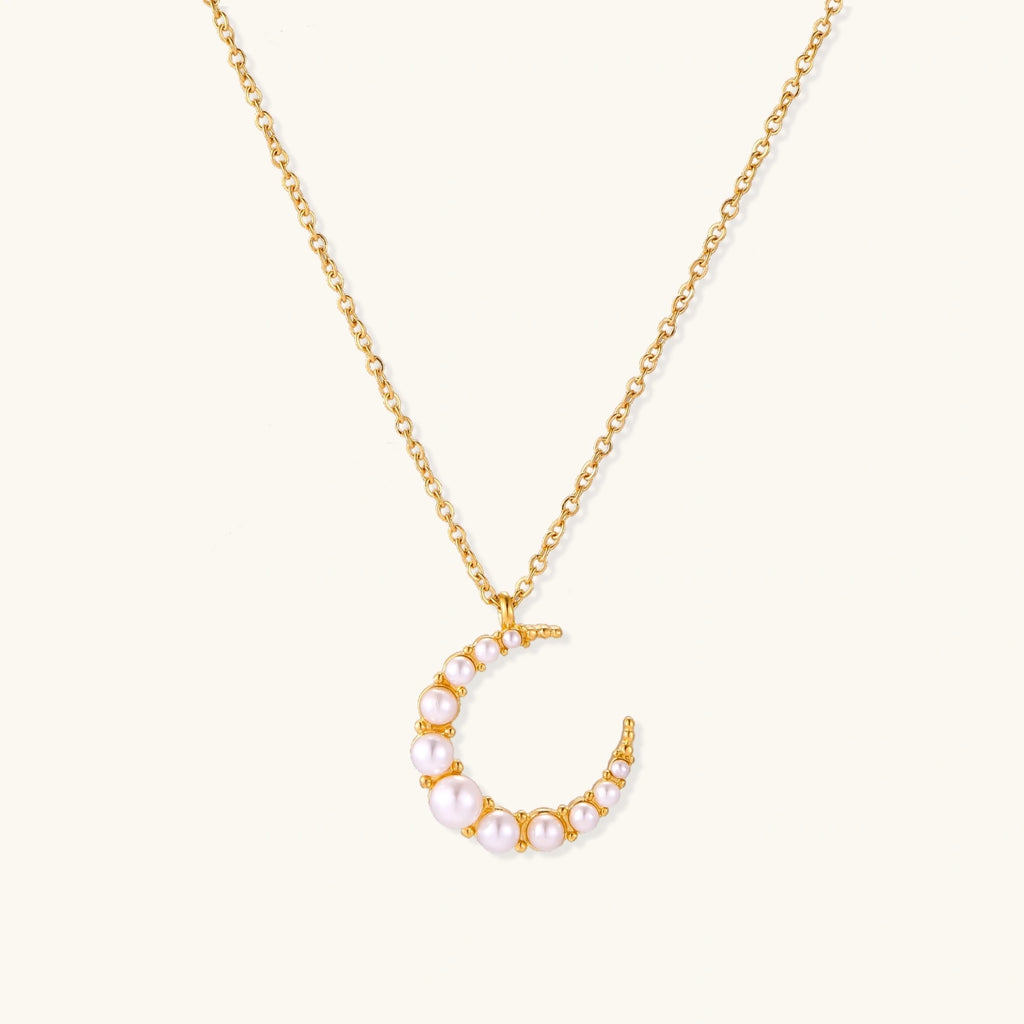 Gold crescent moon pendant with graduated white pearls on delicate chain against cream background