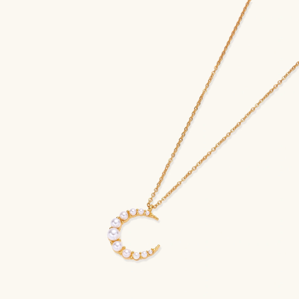 Gold crescent moon pendant with graduated white pearls on delicate chain against cream background