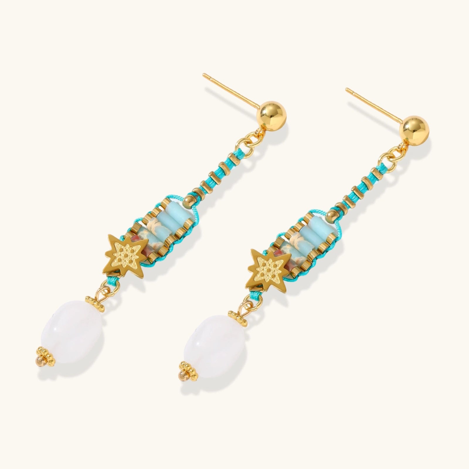 Gold-plated dangle earrings with turquoise threading, star charms, and white pearl beads against cream background.